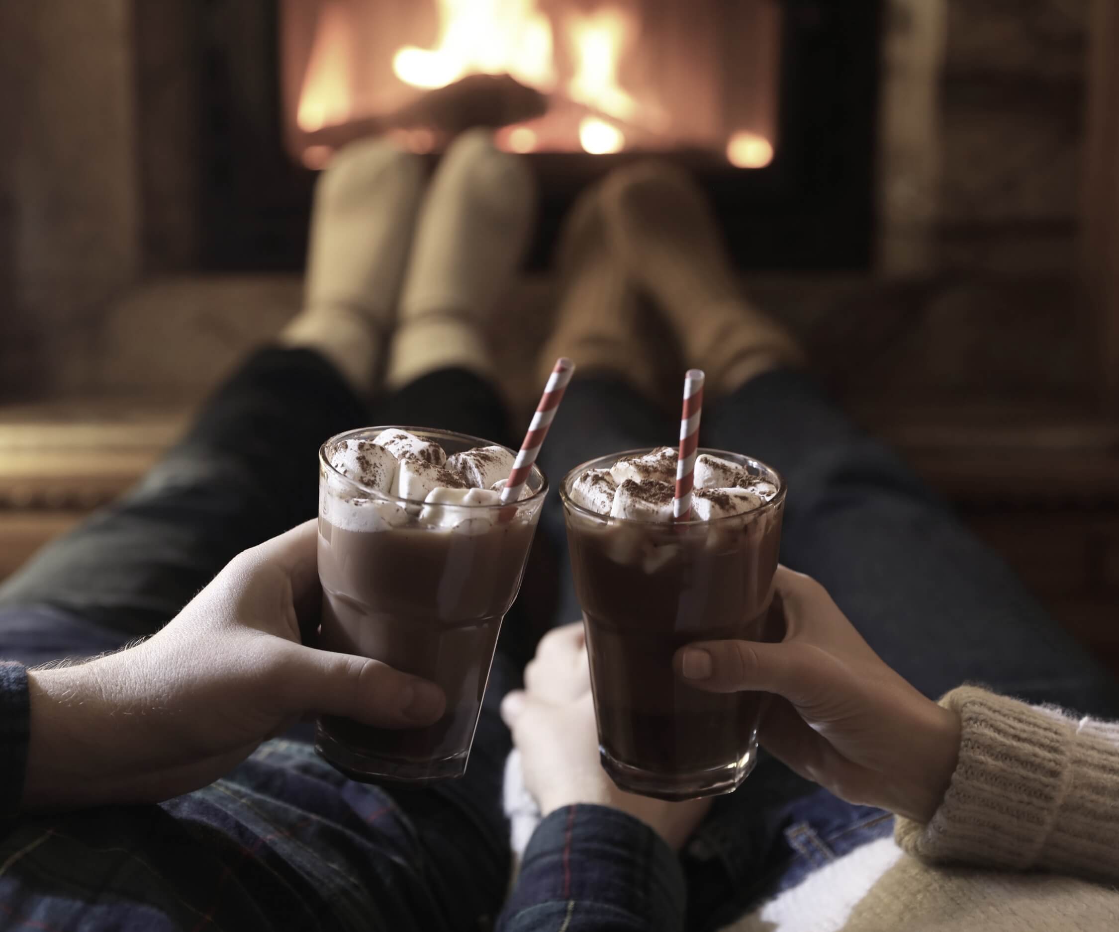 Para siedzi razem przed kominkiem i trzyma w dłoniach kubki gorącej czekolady podczas domowej randki.