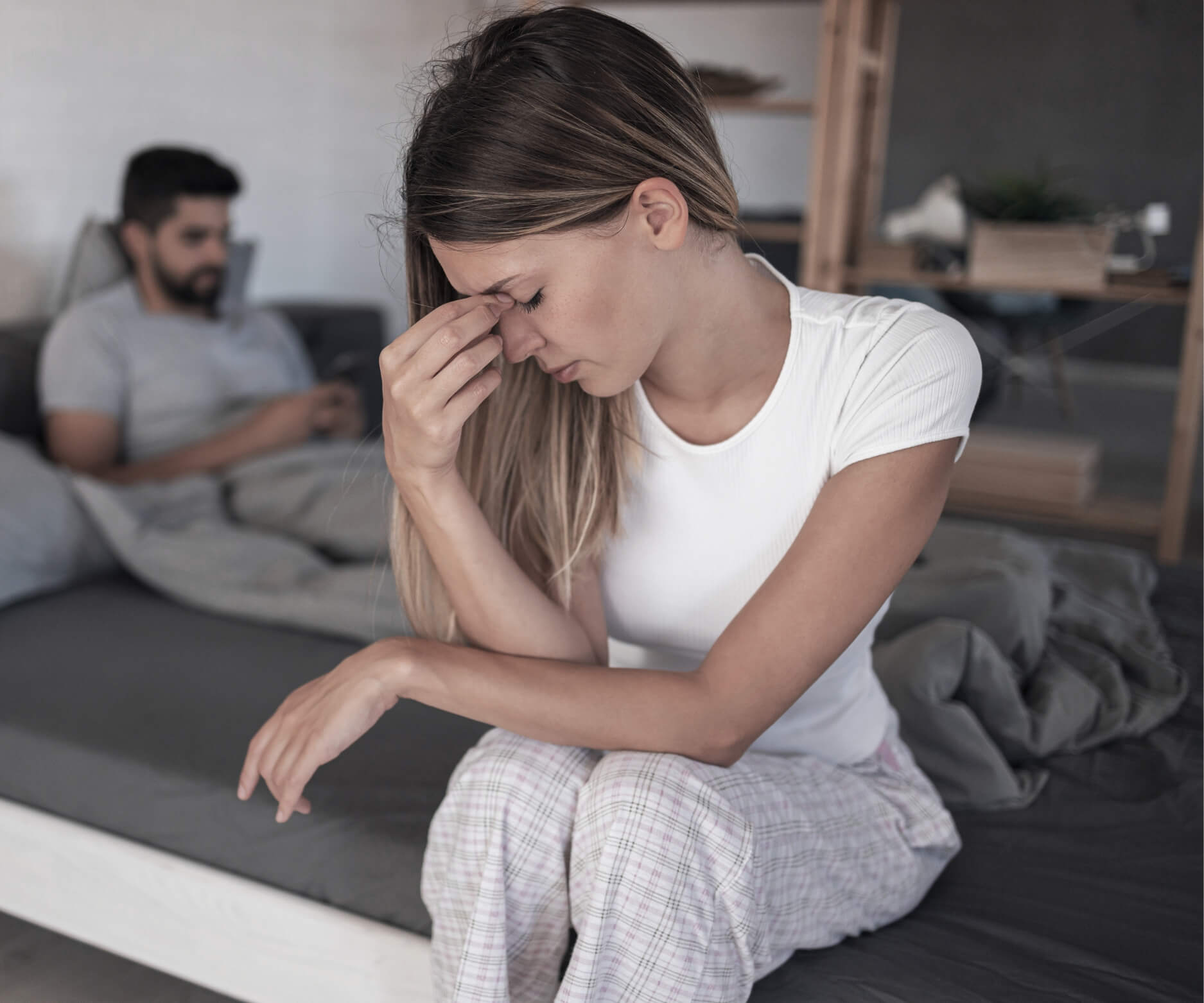 Gaslighting, kontrola i „miłość” – jak rozpoznać przemoc emocjonalną? Na zdjęciu kobieta siedzi zmartwiona na łóżku, w oddali widać mężczyznę, który scrolluje telefon, nie zwracając na nią uwagi.